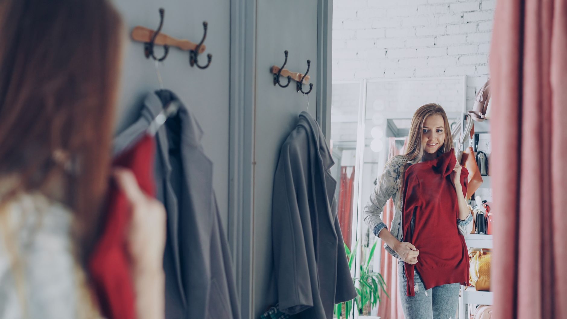 Woman in red shirt and cardigan looking in mirror in home entryway with coats on wall hooks
