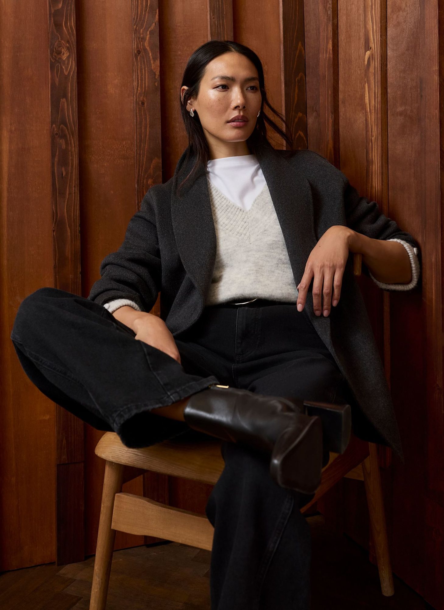 Woman seated on wooden chair wearing charcoal blazer, cream sweater, and black jeans with boots
