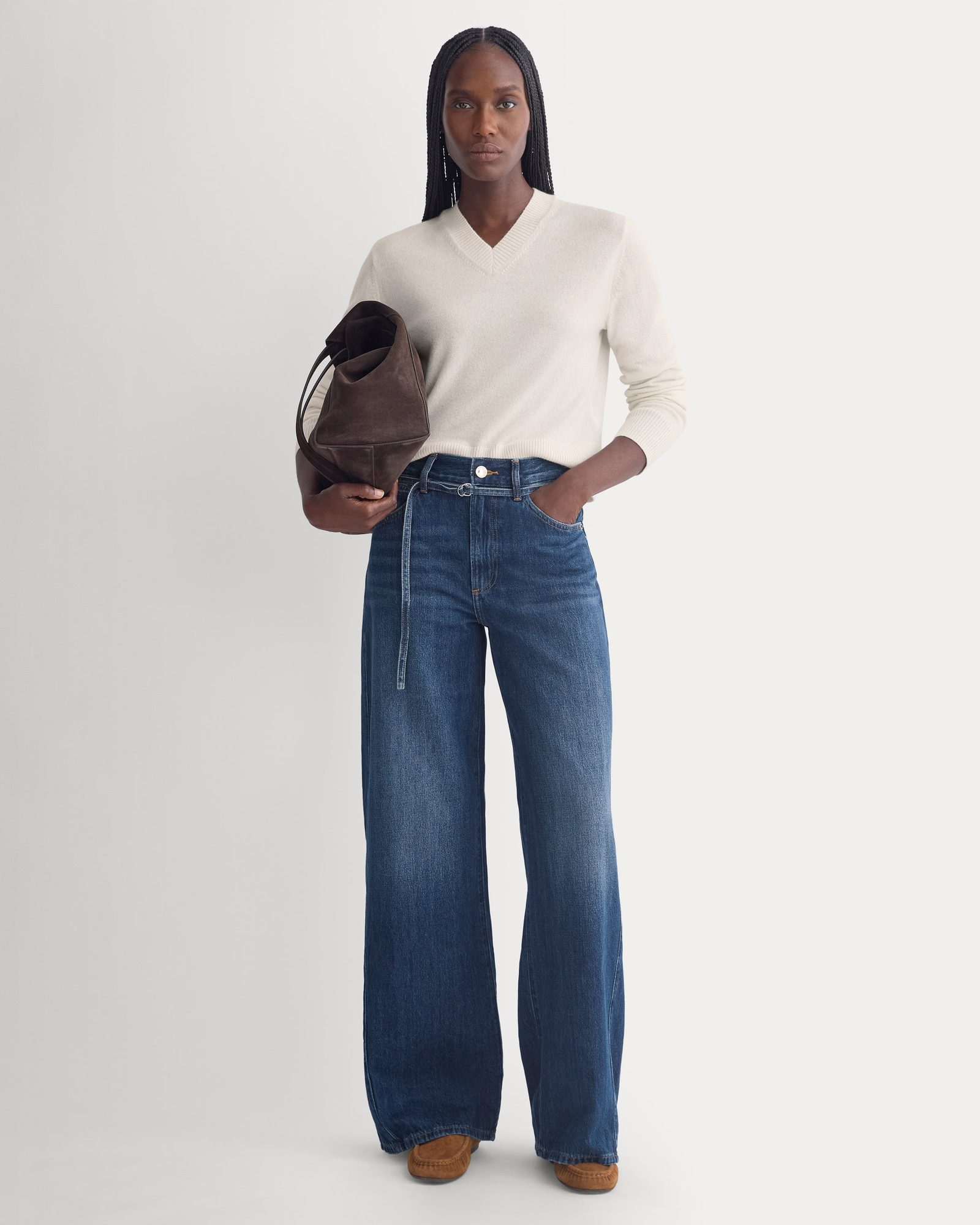 Woman wearing cream v-neck sweater, wide-leg dark blue jeans, and brown suede shoulder bag