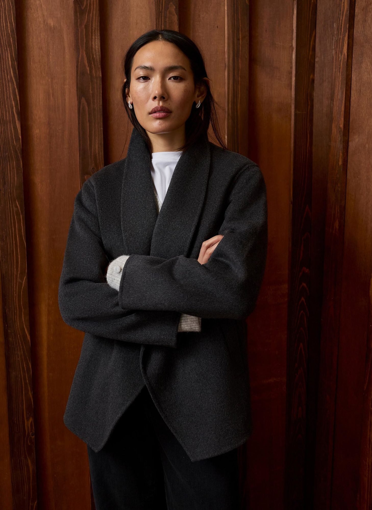 Woman standing with arms crossed wearing charcoal blazer and white shirt against wooden backdrop