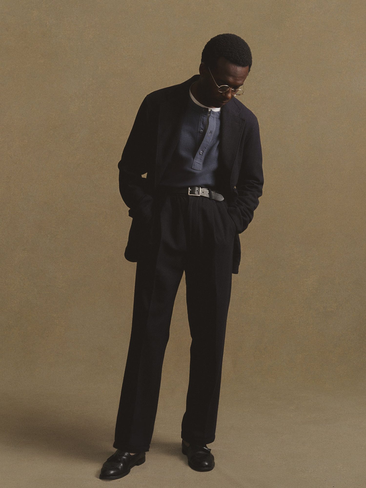 Man in a black blazer layered over a blue henley shirt and black trousers