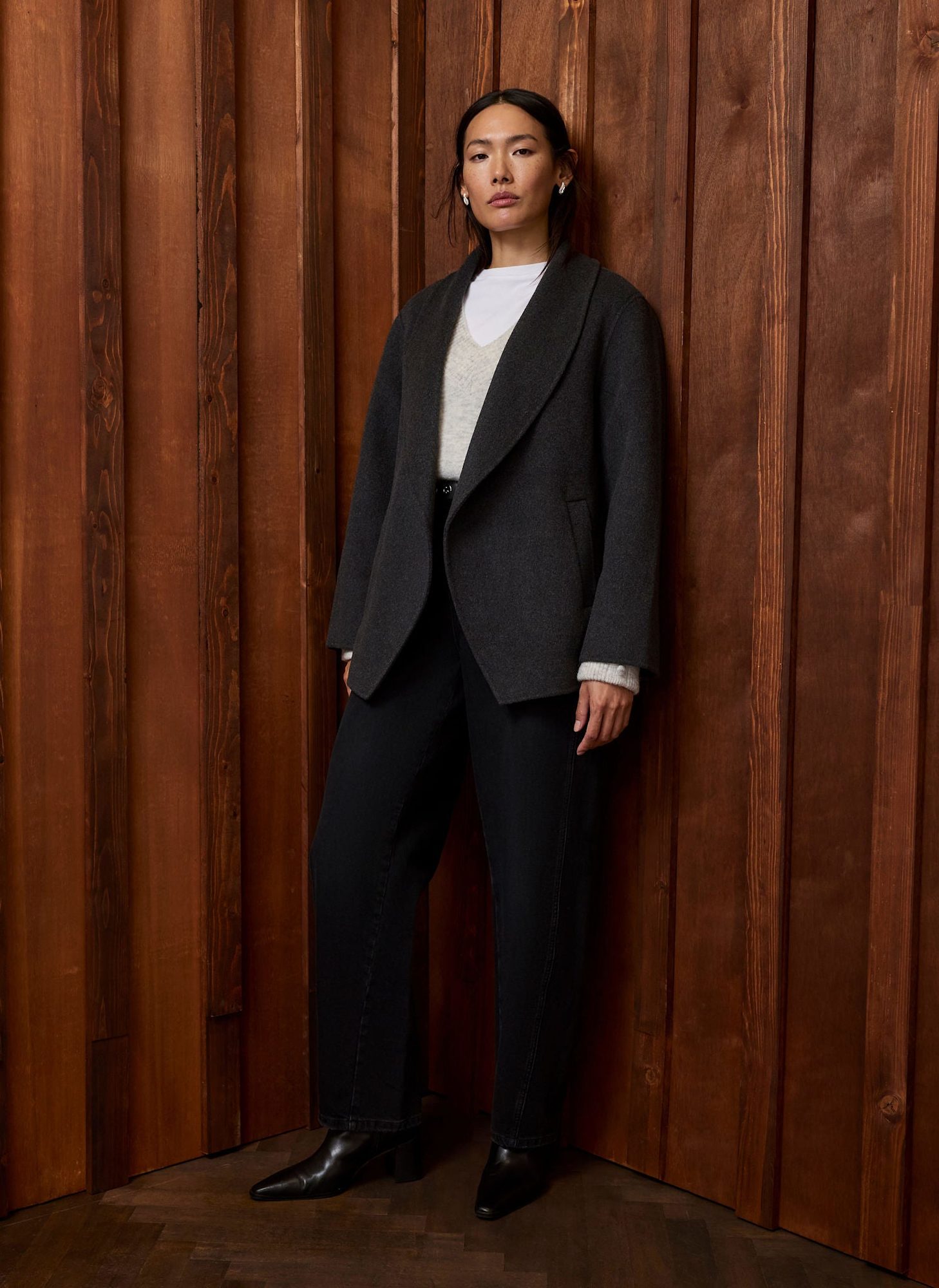 Woman standing in neutral pose wearing charcoal blazer with button detail, white shirt, and black trousers